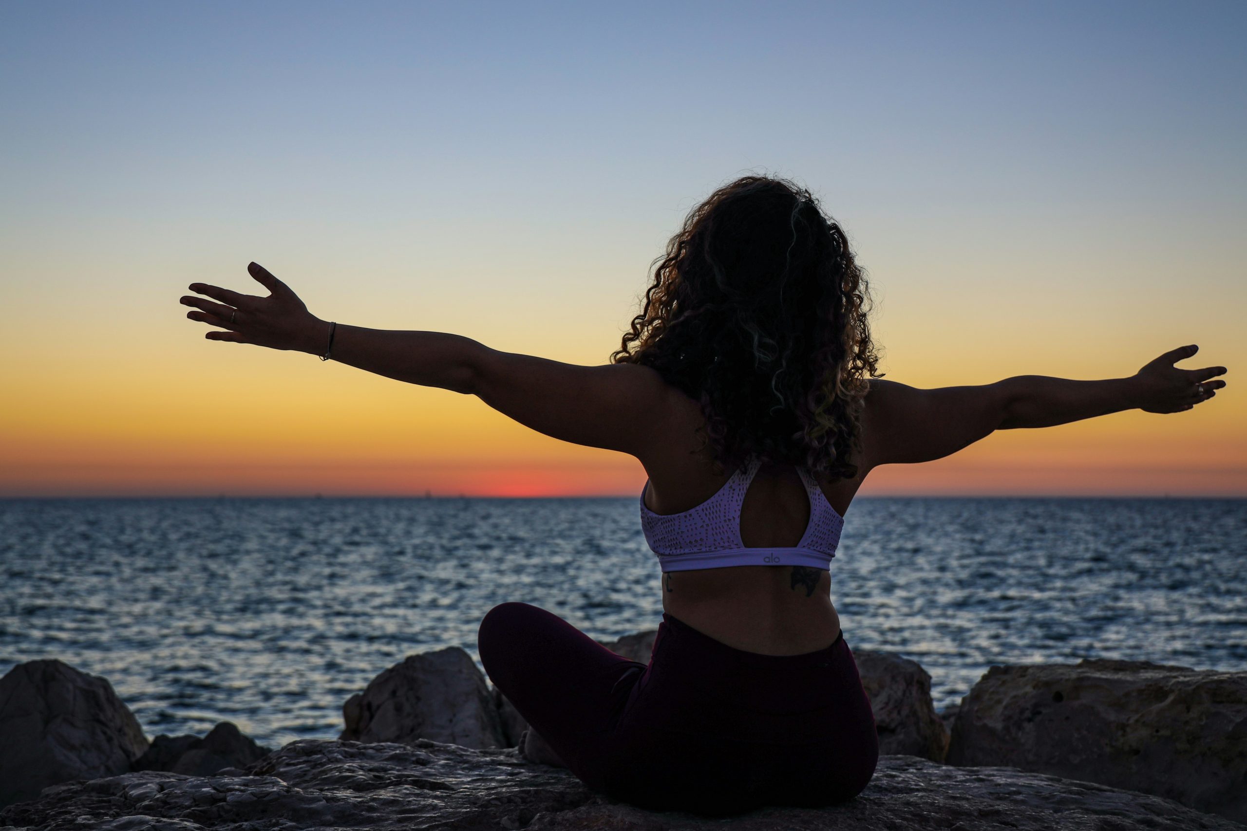 Woman in the stages of menopause sitting on a rock with her legs crossed, arms raised, and looking out at the water.