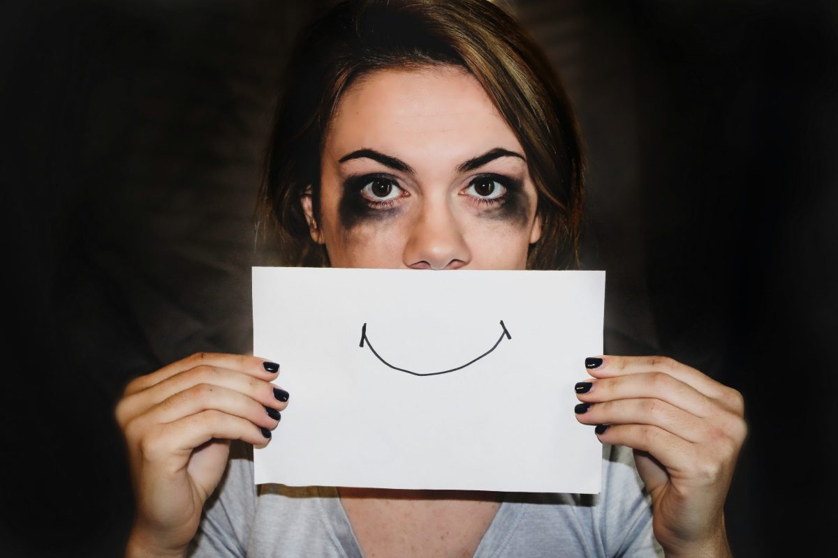 Woman with smudged makeup holding a smiley face over her mouth, symbolizing the emotional rollercoaster of menopause mood swings.