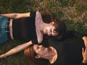 Two women lying head-to-head on the grass, laughing and looking at each other, enjoying a lighthearted moment.