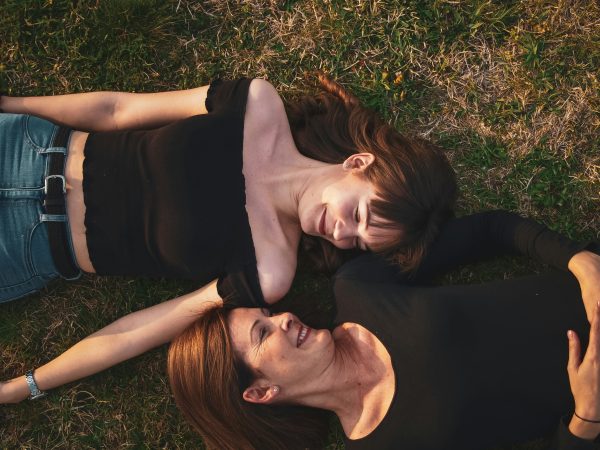 Two women lying head-to-head on the grass, laughing and looking at each other, enjoying a lighthearted moment.