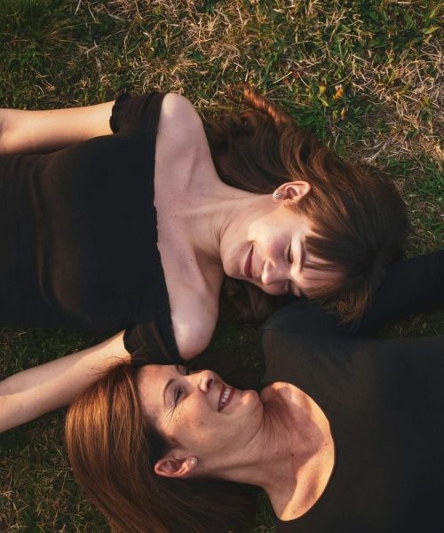 Two women lying head-to-head on the grass, laughing and looking at each other, enjoying a lighthearted moment.