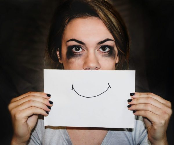 Woman with smudged makeup holding a smiley face over her mouth, symbolizing the emotional rollercoaster of menopause mood swings.